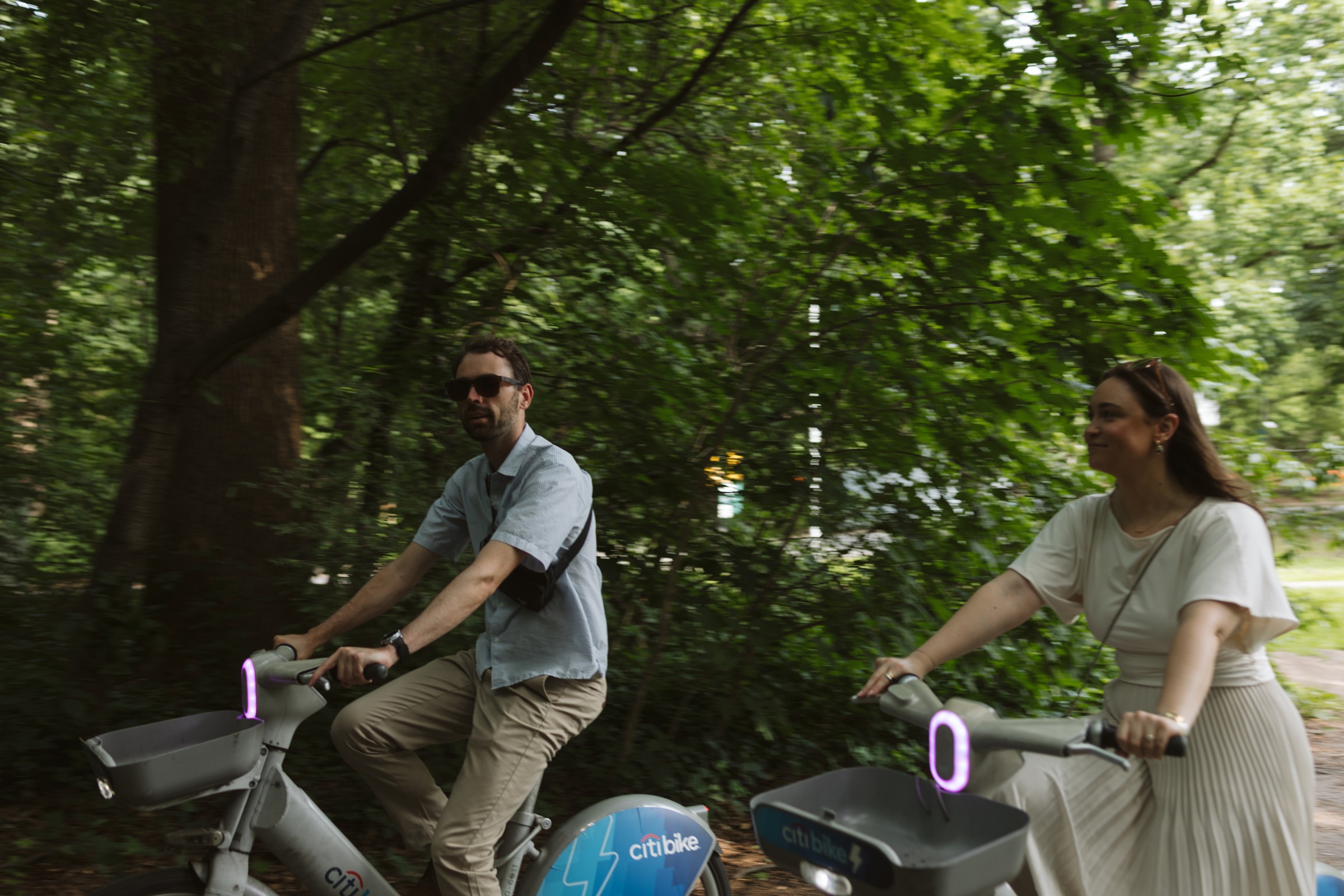 Monika and Haggai riding Citi Bikes side by side down a tree-lined park road.